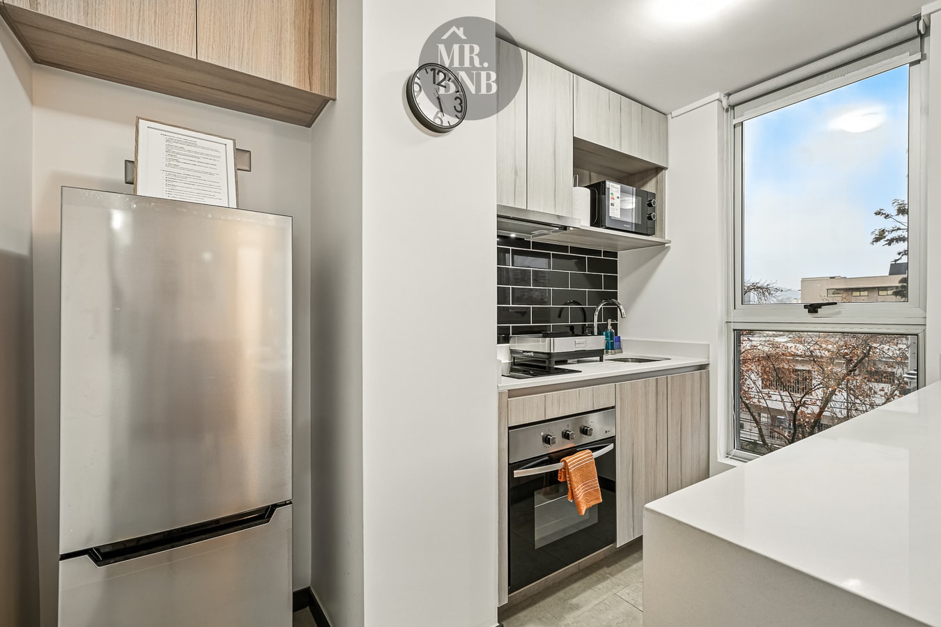 Cocina con refrigerador, horno, muebles claros y ventana con vista a árboles.
