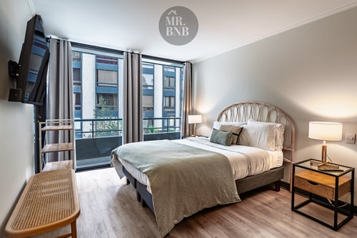 Dormitorio principal con cama, cabecero de mimbre, velador, TV y ventanal hacia balcón con vista a edificios