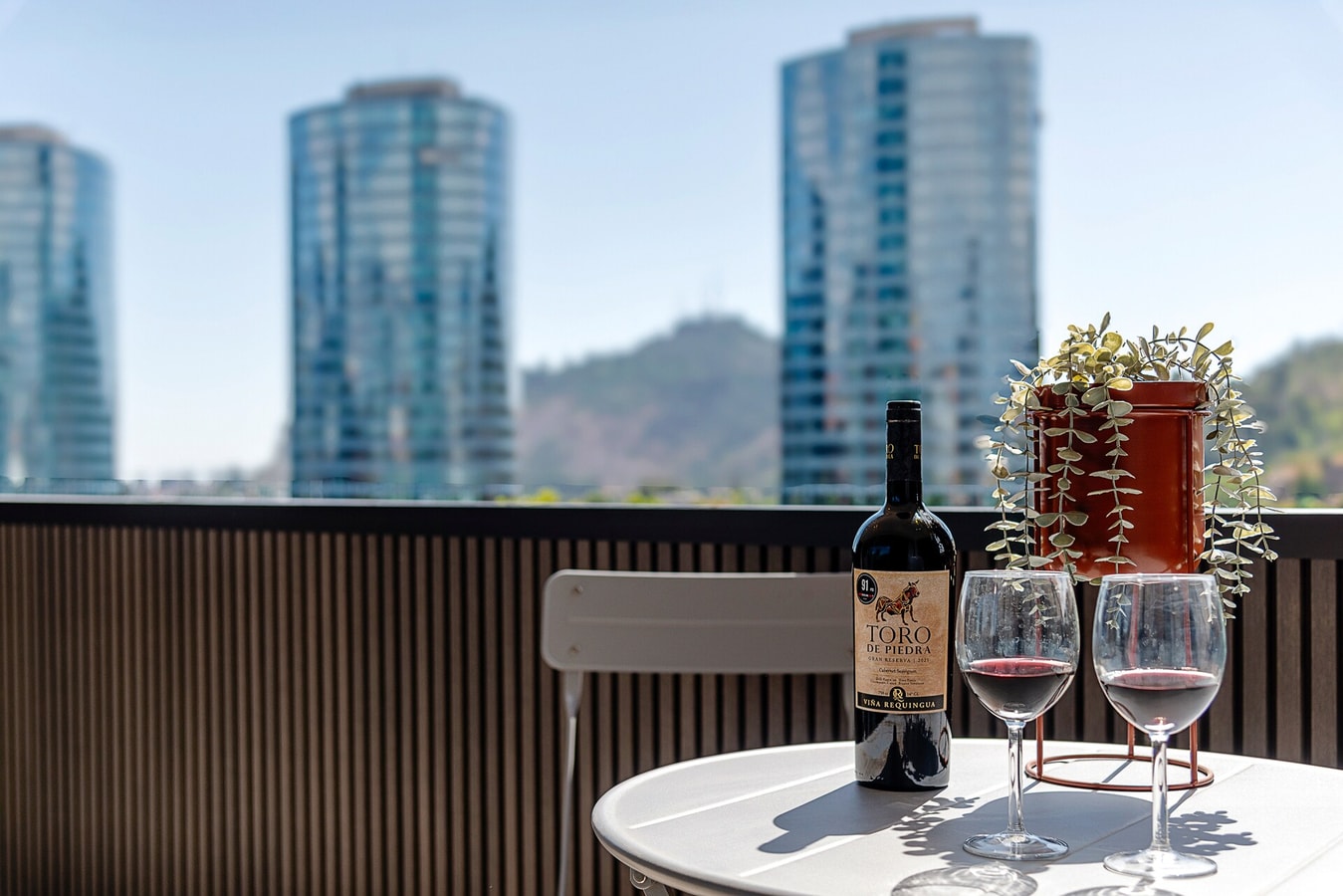 Detalle de la mesa de la terraza con una botella de vino y dos copas servidas, y las torres de vidrio del barrio al fondo