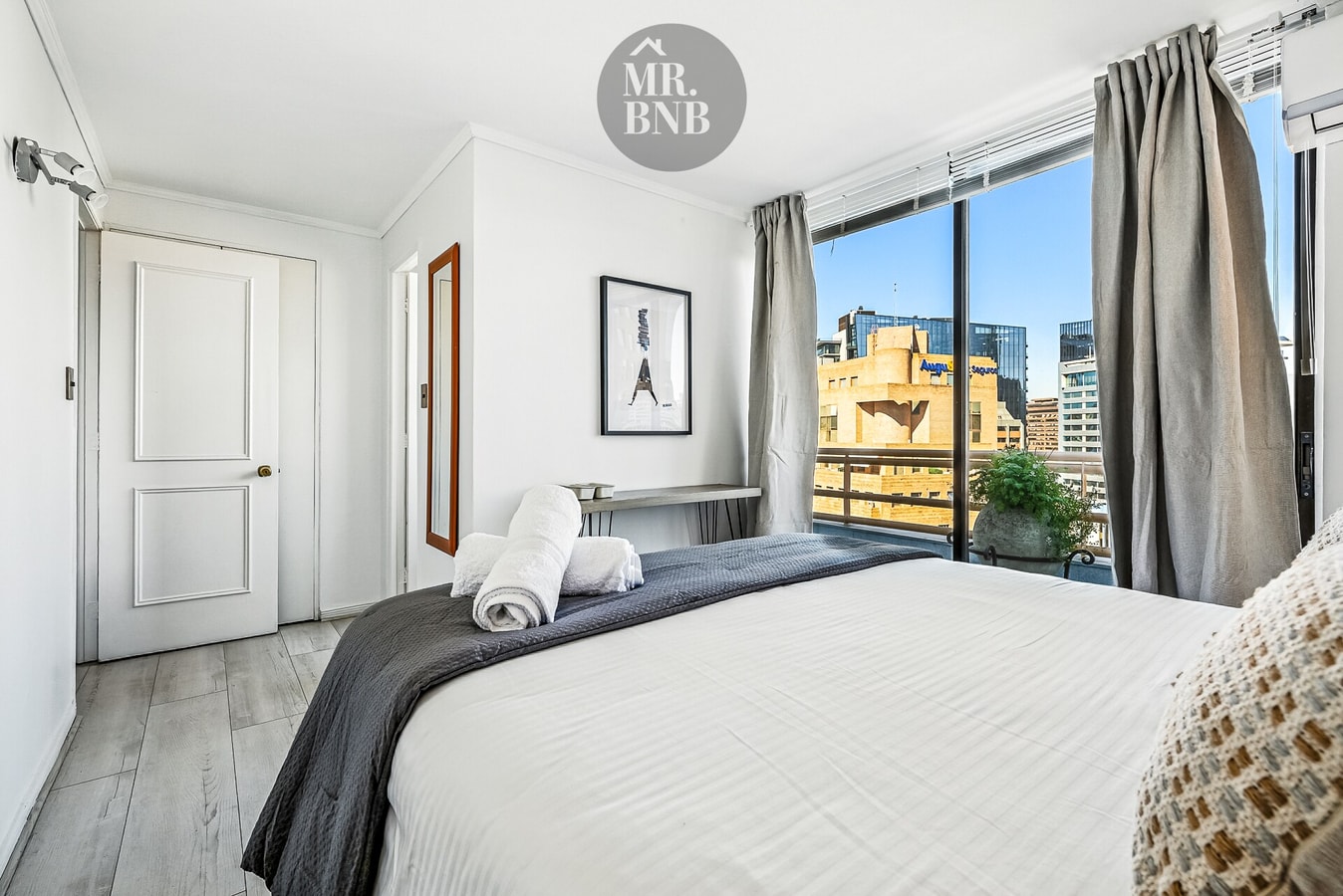 Dormitorio con cama, mesa auxiliar y ventanal con vista a edificios