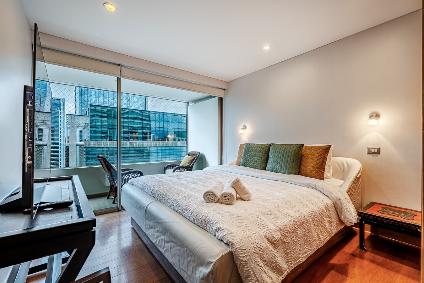 Dormitorio con cama, ventanal con vista a edificios y mesa de living.
