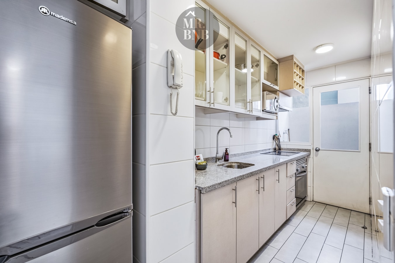 Cocina con refrigerador, muebles blancos y puerta hacia el pasillo.