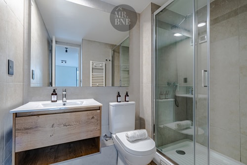 Baño con mueble de madera, doble lavamanos, espejo, inodoro y ducha de vidrio