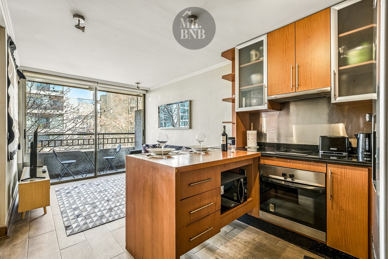 Cocina con muebles de madera, balcón al fondo con mesa y sillas, microondas