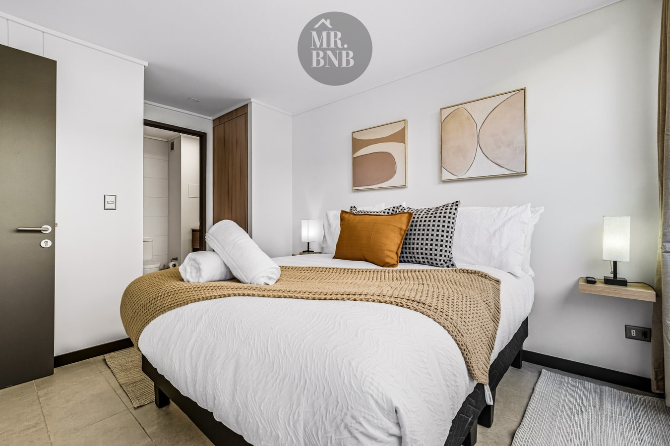 Dormitorio con cama de dos plazas, manta tejida, cojines naranjos y dos láminas sobre el respaldo