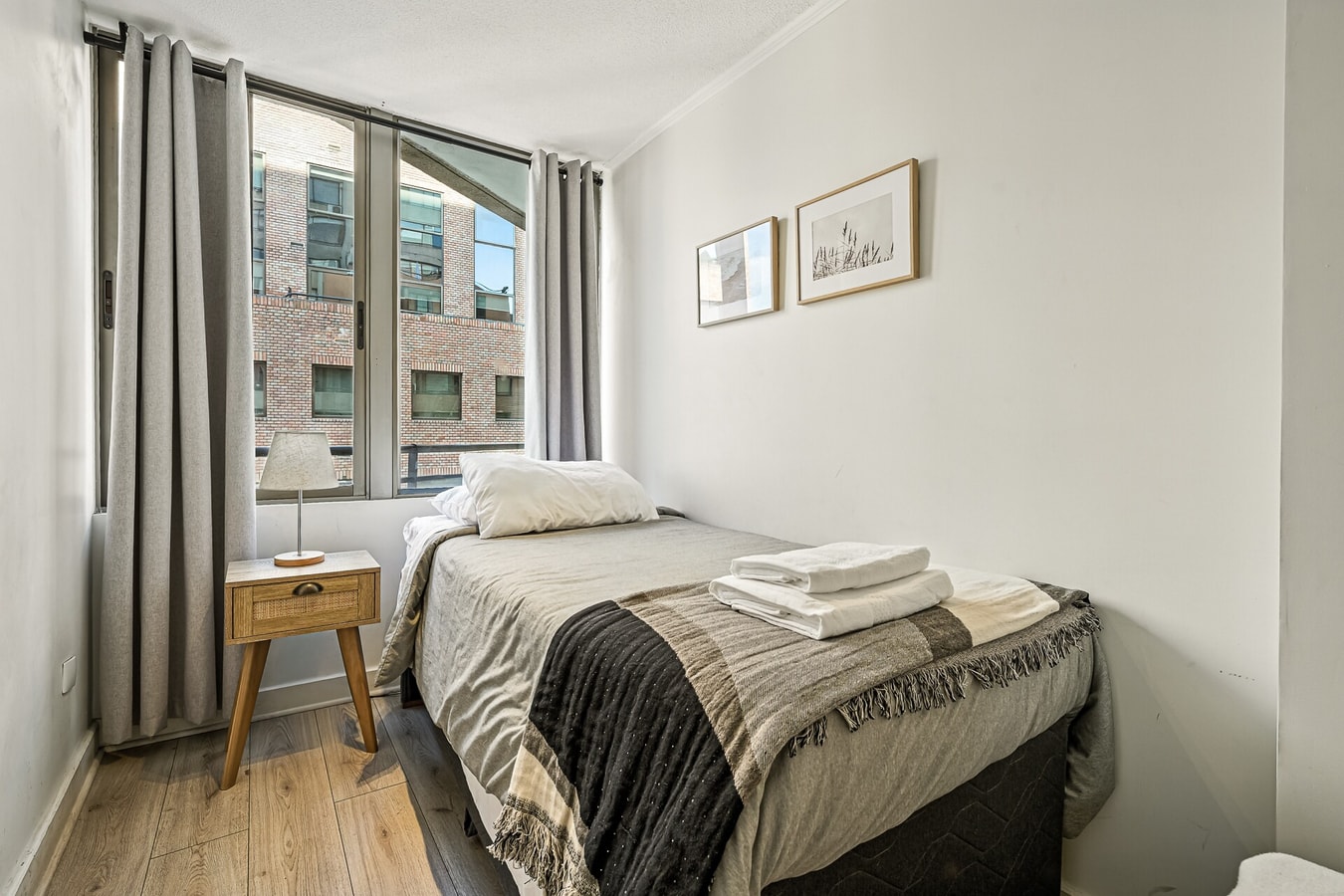 Dormitorio con cama, veladores y ventanal hacia edificios vecinos