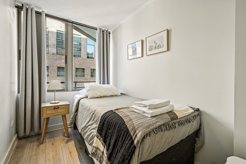 Dormitorio con cama, veladores y ventanal hacia edificios vecinos