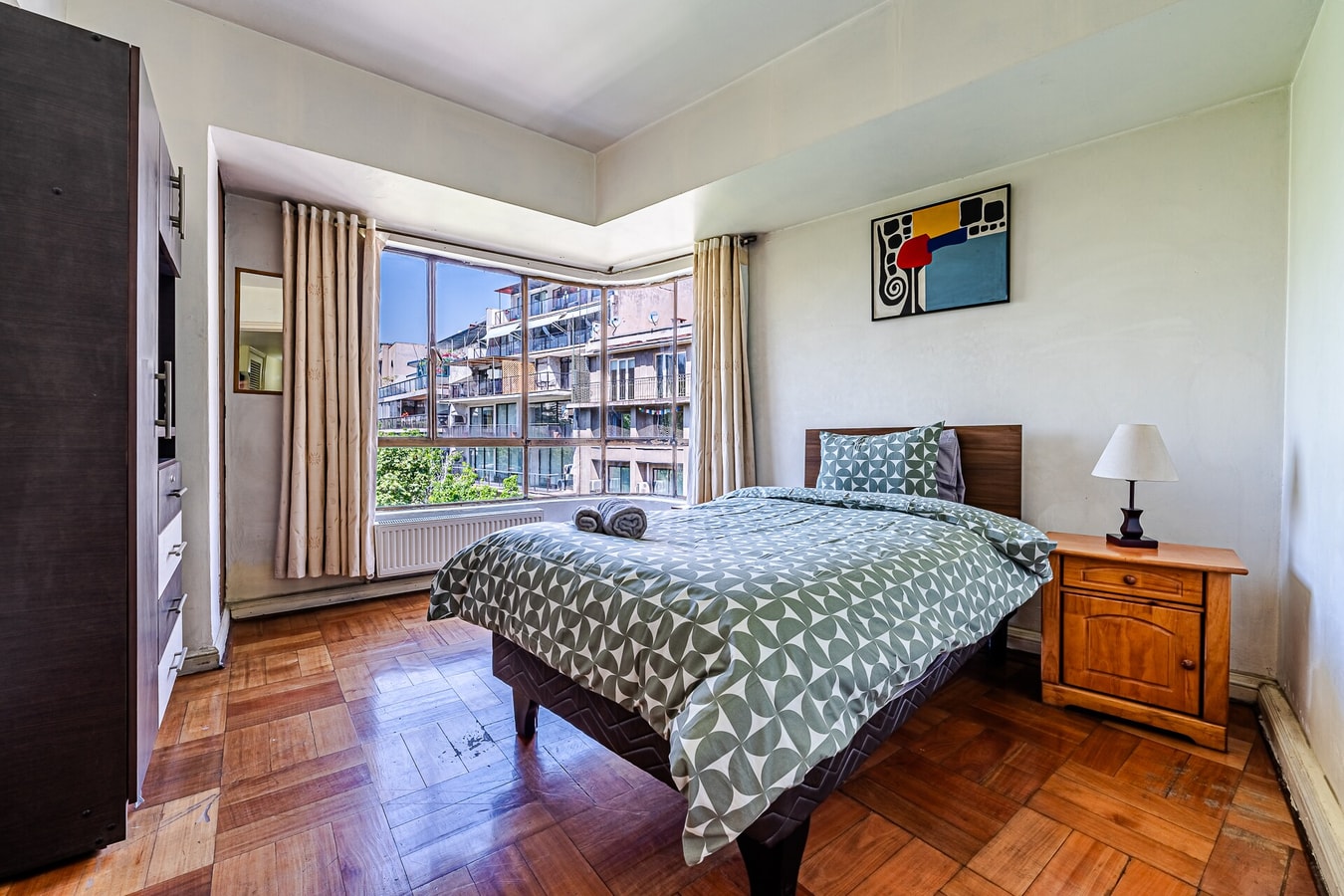 Dormitorio con cama matrimonial, veladores, cuadro y ventana con vista a edificios
