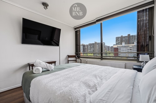 Dormitorio con cama, radiador y ventanal con vista a edificios.