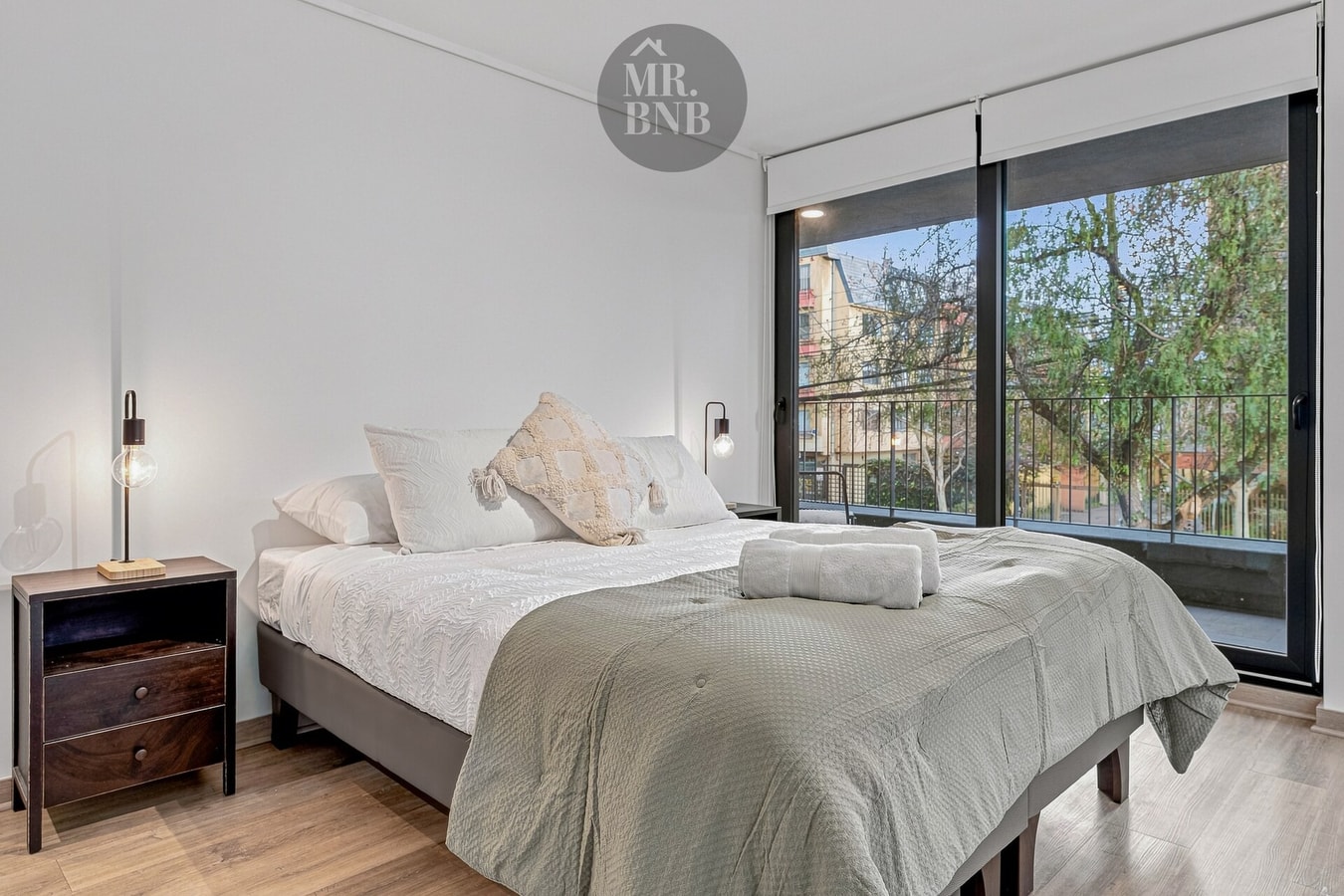 Dormitorio con cama doble, velador de madera y ventana hacia árboles