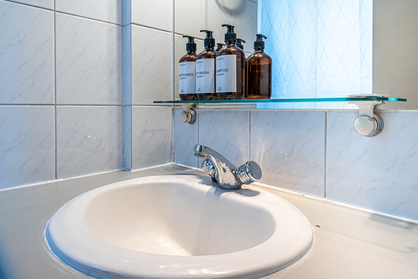 Detalle de lavamanos de baño con amenities en frascos sobre repisa de vidrio.