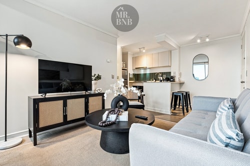 Living con sofá, mueble de TV, mesa de centro y cocina con barra y taburetes al fondo