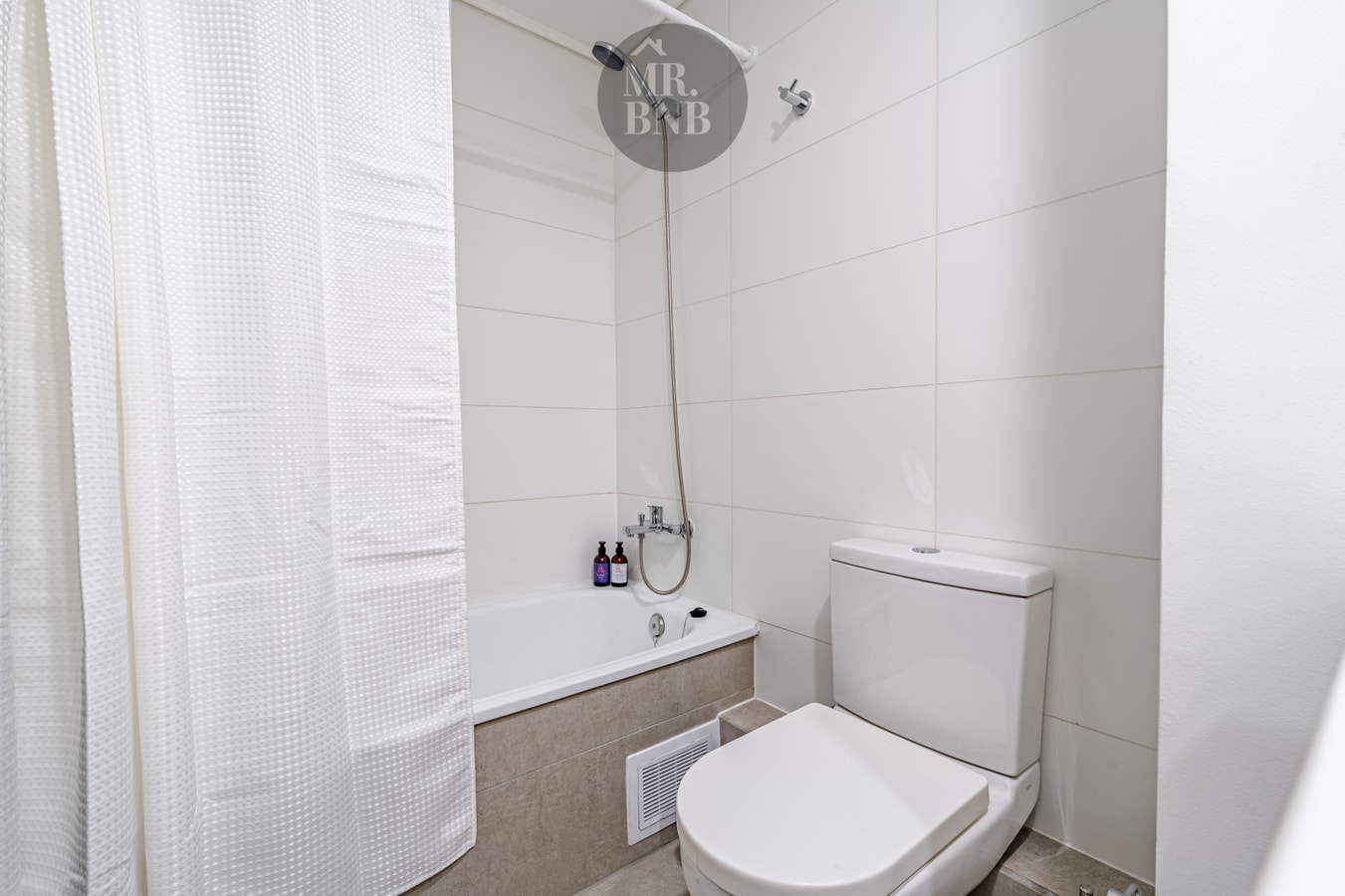 Baño con tina, cortina blanca, ducha teléfono e inodoro, revestido en cerámica blanca