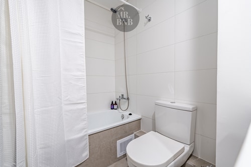 Baño con tina, cortina blanca, ducha teléfono e inodoro, revestido en cerámica blanca