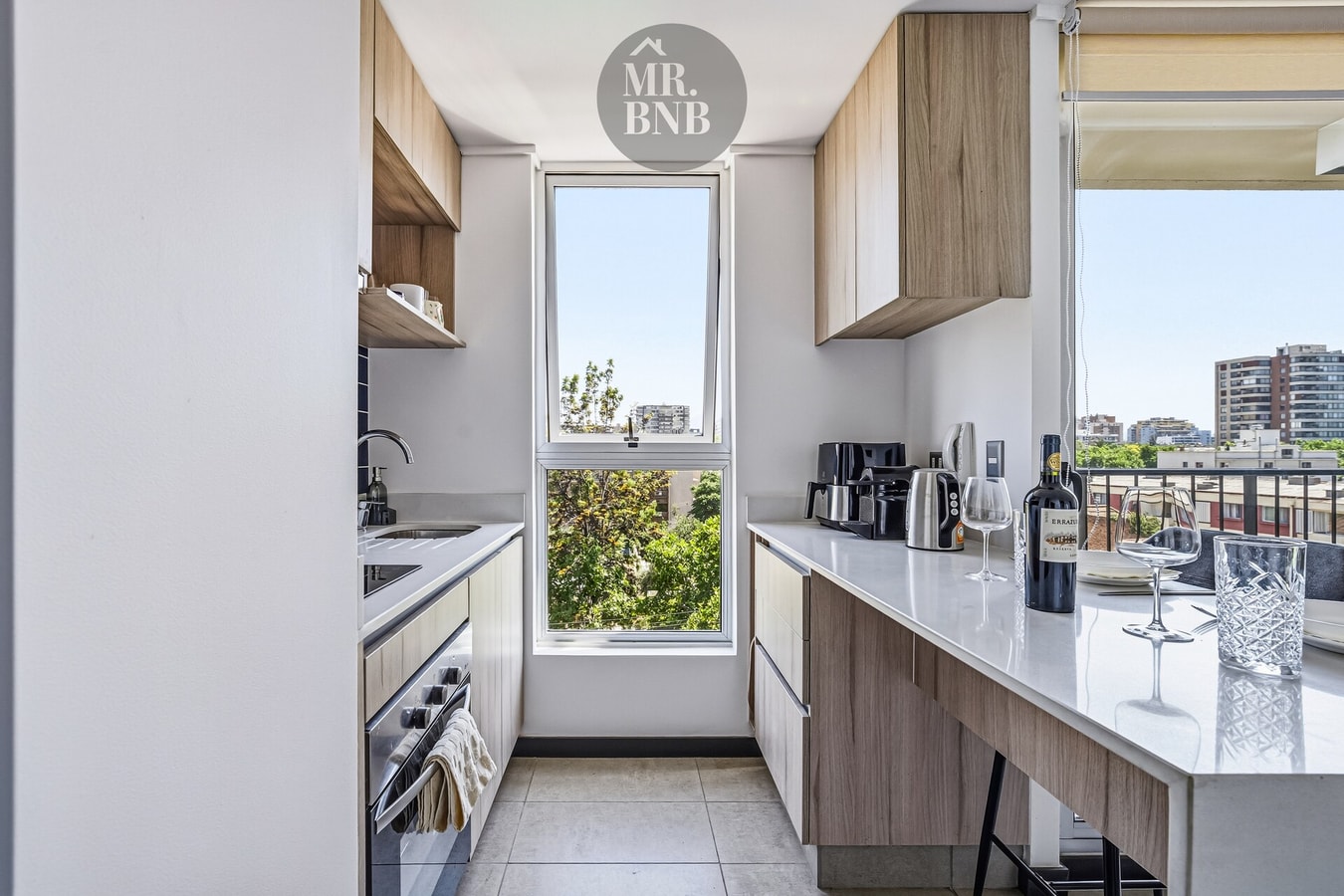 Cocina en línea con muebles claros, horno, ventana y vista hacia árboles