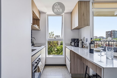 Cocina en línea con muebles claros, horno, ventana y vista hacia árboles