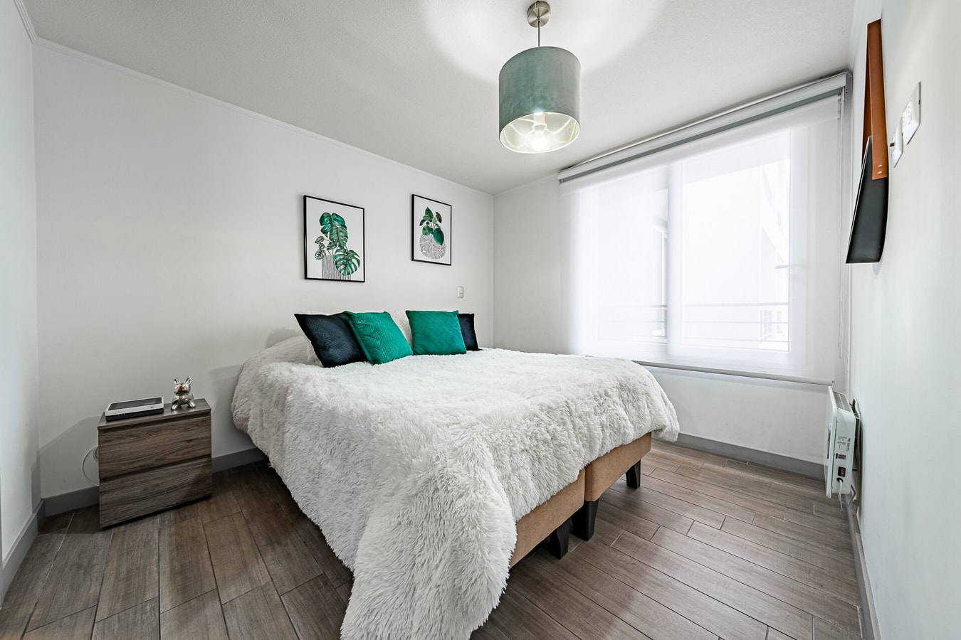 Dormitorio con cama de cojines verdes, ventana con cortina y calefactor eléctrico junto a la cama