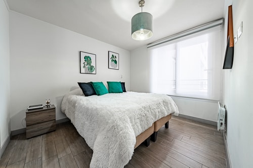 Dormitorio con cama de cojines verdes, ventana con cortina y calefactor eléctrico junto a la cama