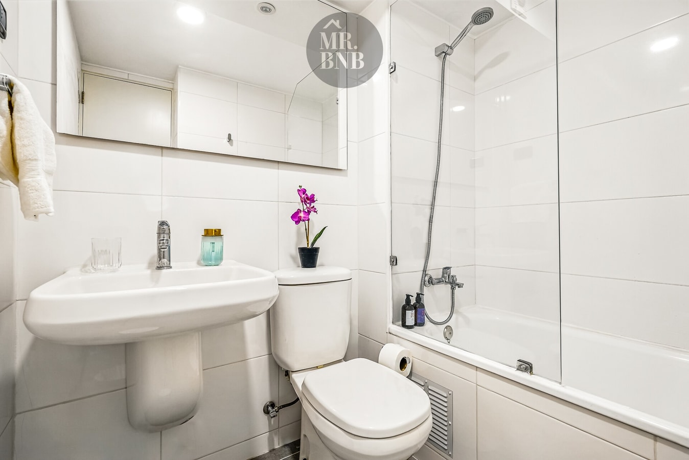 Baño con lavamanos, inodoro, tina con ducha y detalle de flor sobre el mueble.