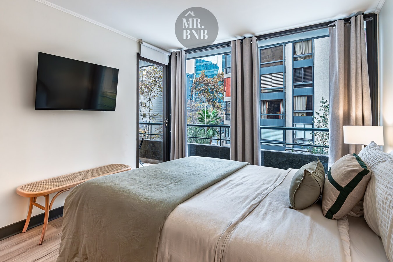 Dormitorio principal con cama, TV en la pared, ventanales hacia balcón y vista a edificios