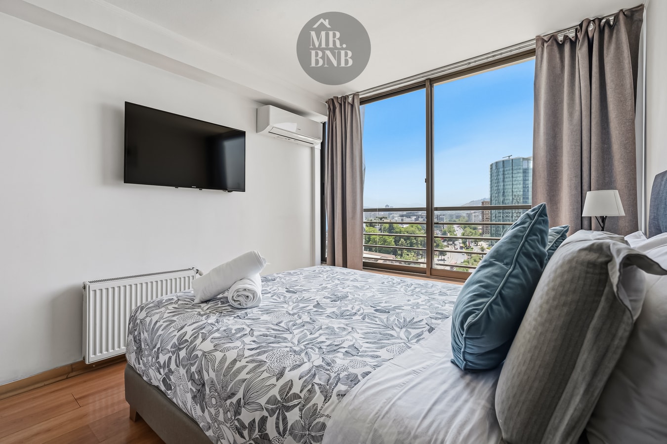 Dormitorio con cama, televisor en la pared y ventanal con vista a la ciudad.