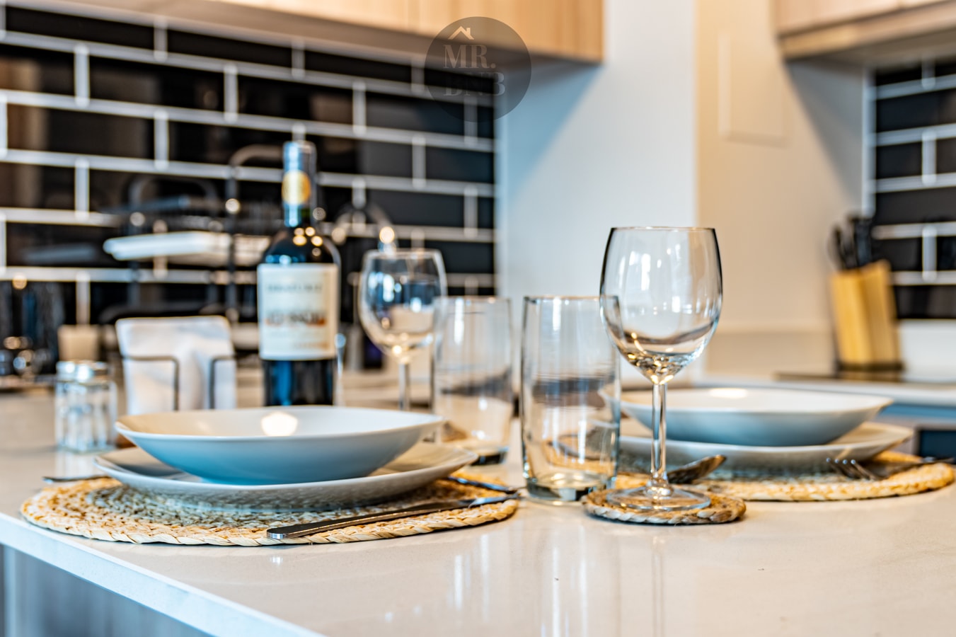 Detalle de mesa puesta sobre el mesón: platos hondos, copas, vasos e individuales de fibra, con una botella de vino al fondo