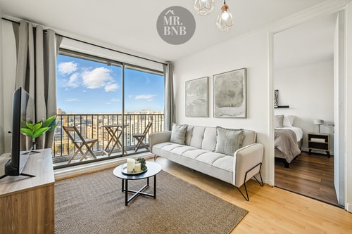 Living con sofá blanco, mesa de centro, alfombra y ventanal hacia la ciudad, con vista al dormitorio