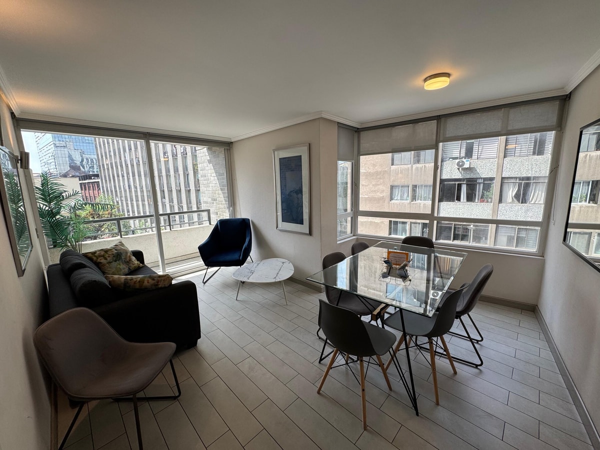 Living comedor con sofá gris, poltronas, mesa de comedor de vidrio con seis sillas y ventanales a la ciudad