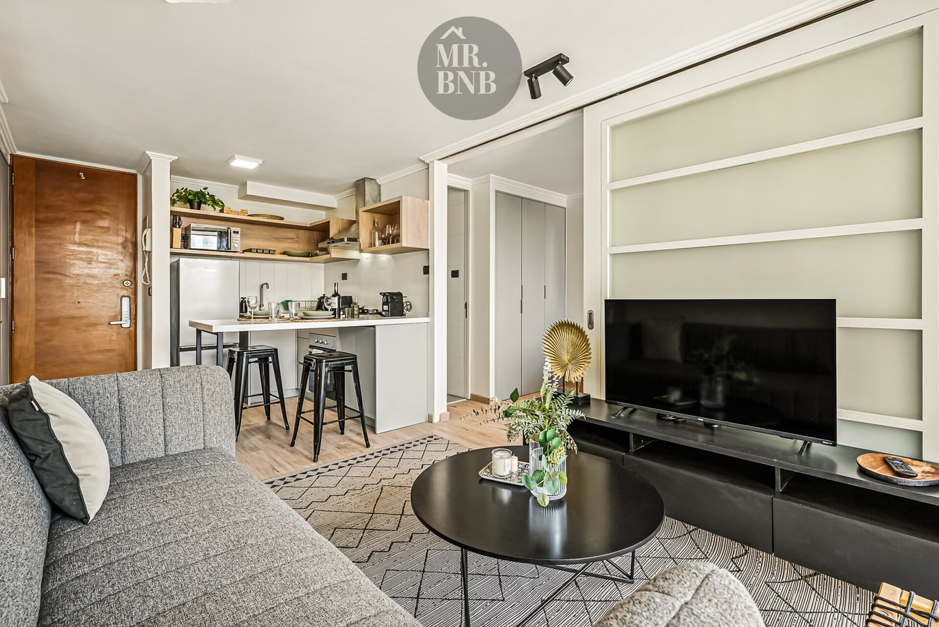 Living con sofá gris, mesa de centro, mueble para televisor y vista hacia la cocina.