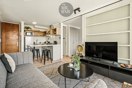 Living con sofá gris, mesa de centro, mueble para televisor y vista hacia la cocina.