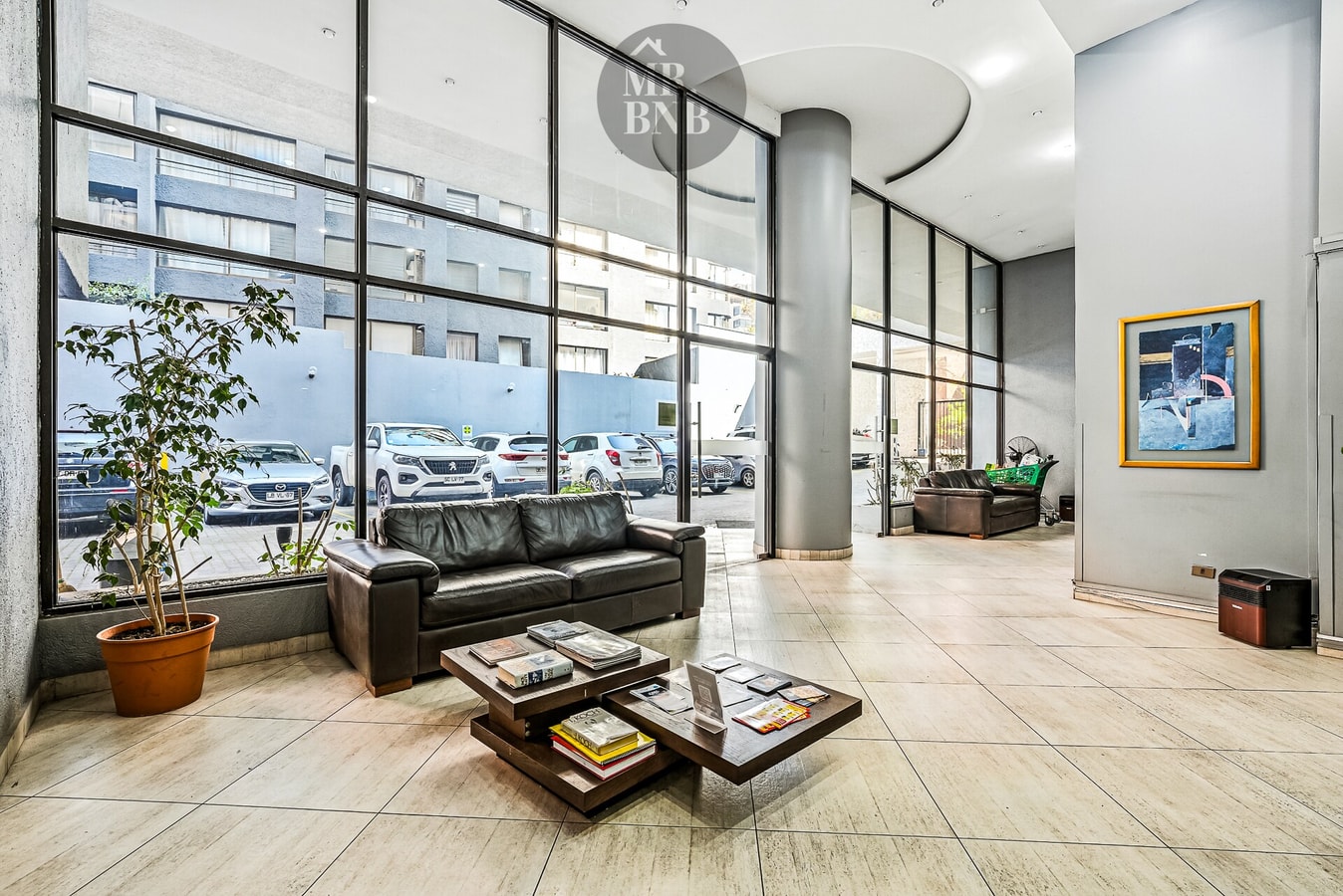 Lobby del edificio con sofá de cuero, mesa de centro y ventanales hacia el estacionamiento