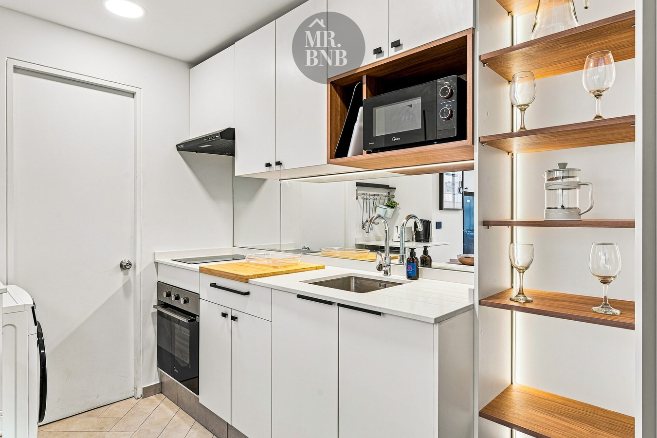 Cocina blanca con horno, microondas y repisas de madera con copas.