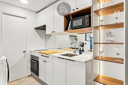 Cocina blanca con horno, microondas y repisas de madera con copas.