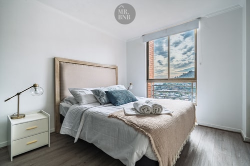 Dormitorio con cama matrimonial, respaldo tapizado y ventanal con vista a la ciudad y los cerros