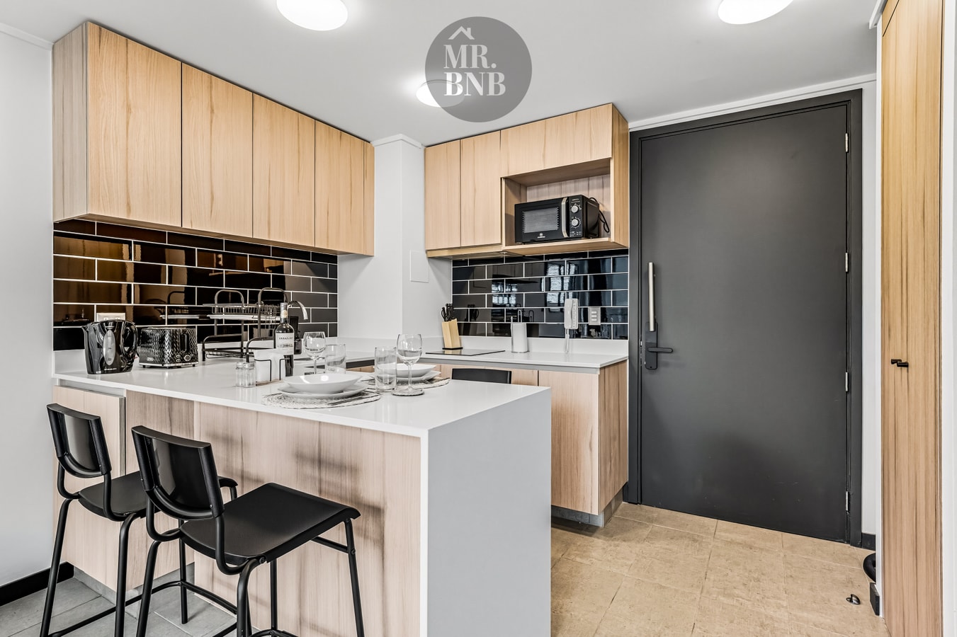 Cocina con barra y dos banquetas negras junto a la puerta de acceso del departamento, muebles de madera clara y microondas empotrado