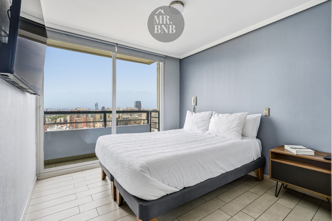 Dormitorio con cama matrimonial, velador de madera y ventanal con vista a la ciudad