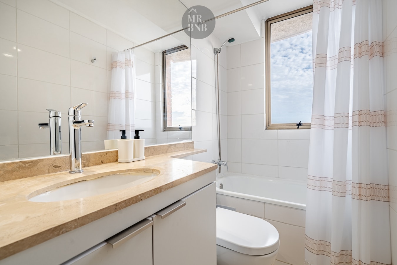 Baño con tina, cortina a rayas y lavamanos de mármol con dos llaves, muy similar al anterior