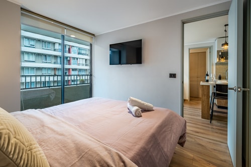 Dormitorio con cama, puerta corredera de vidrio hacia el pasillo y televisor en la pared.