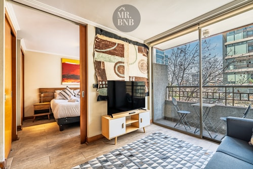 Dormitorio con cama y TV, con vista al living con sofá y ventanal hacia balcón con edificios