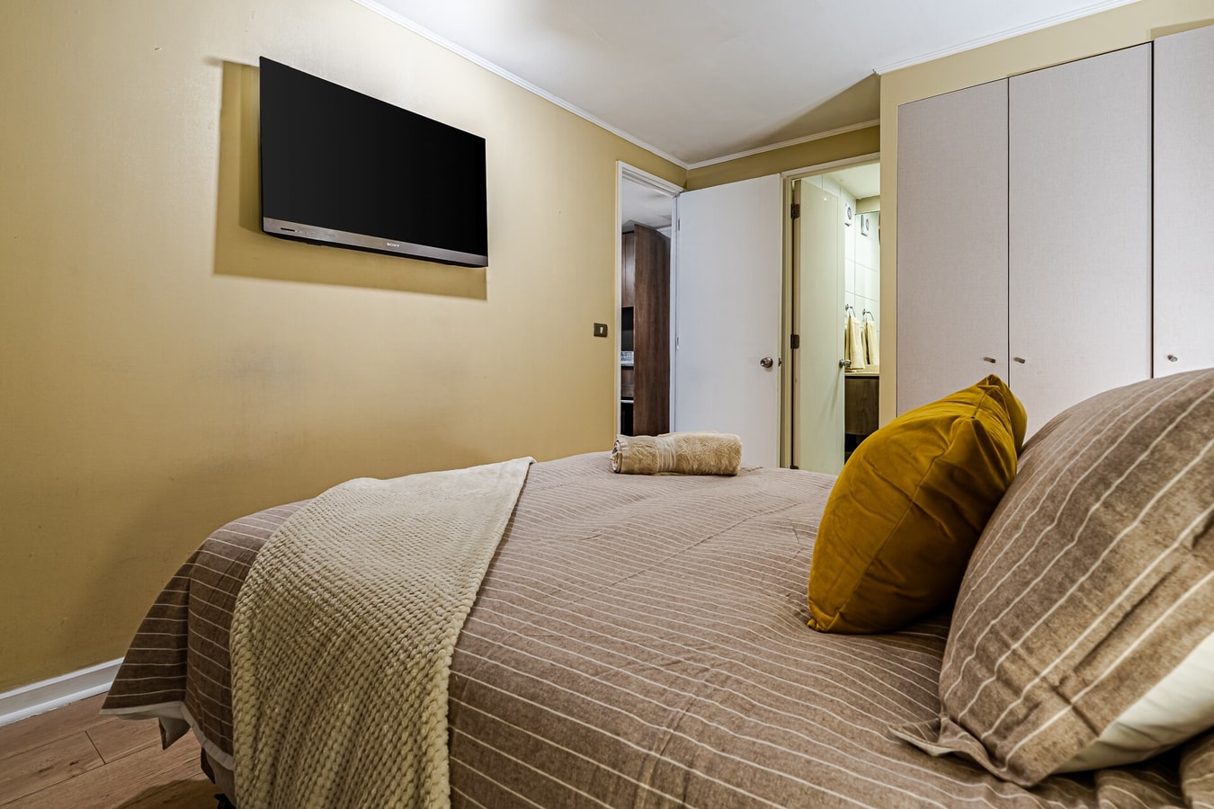 Dormitorio en suite con TV, cama y cojines mostaza