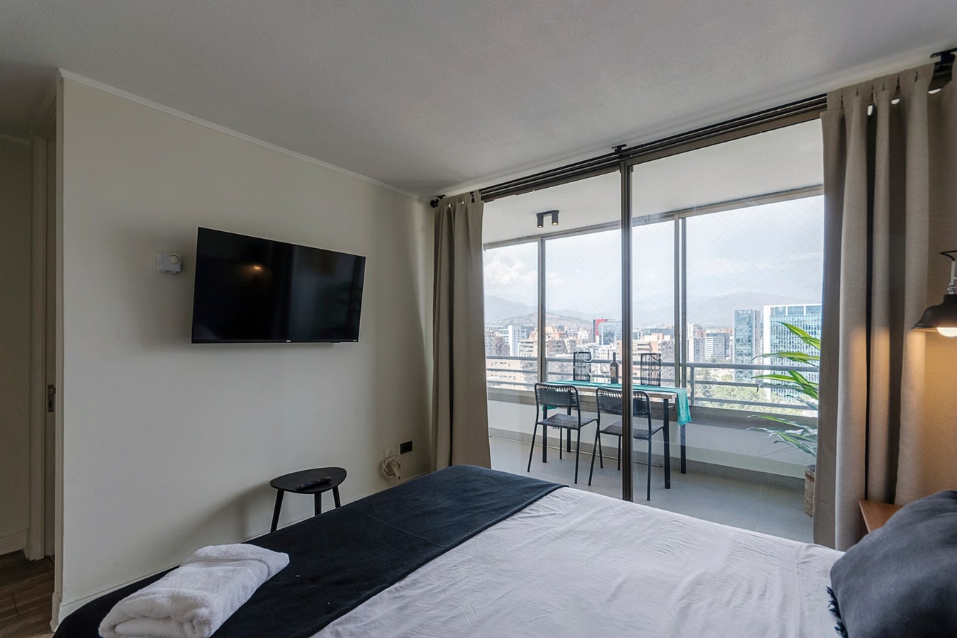 Dormitorio con cama, TV en la pared, ventanal hacia terraza con mesa y sillas, vista a la ciudad