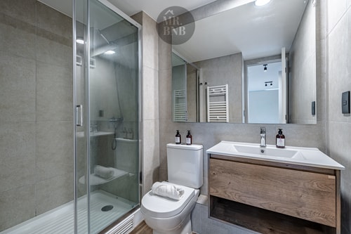 Baño con ducha de vidrio, mueble de madera con lavamanos y toallas blancas dispuestas