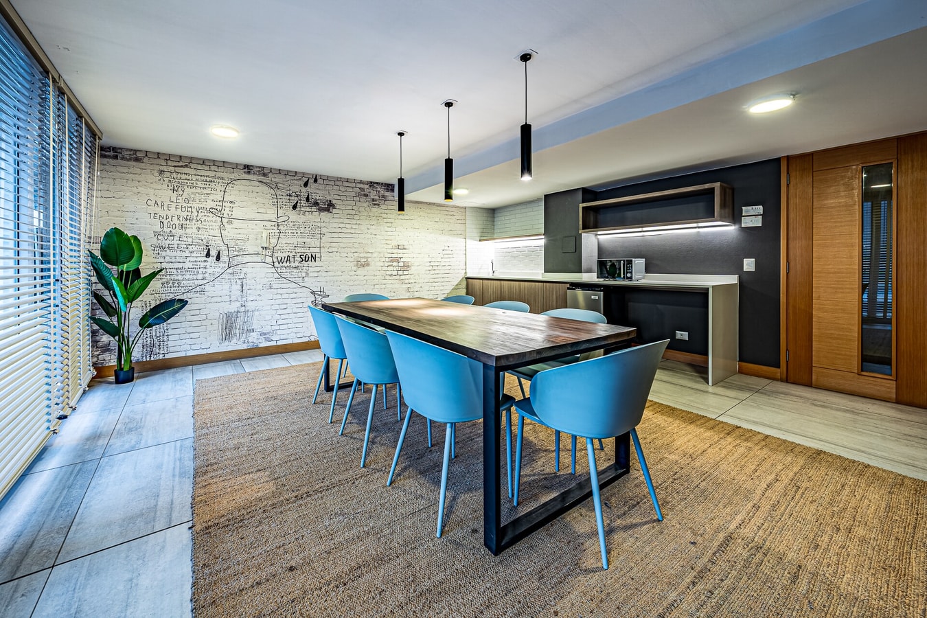 Salón común del edificio con mesa de comedor larga, sillas azules tapizadas y cocina integrada al fondo
