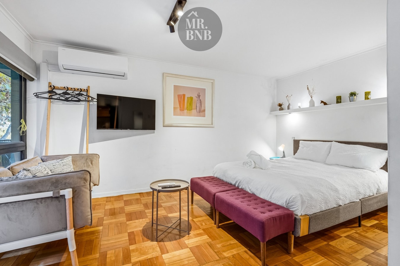 Dormitorio de suite con cama, banca color vino, sillón y TV
