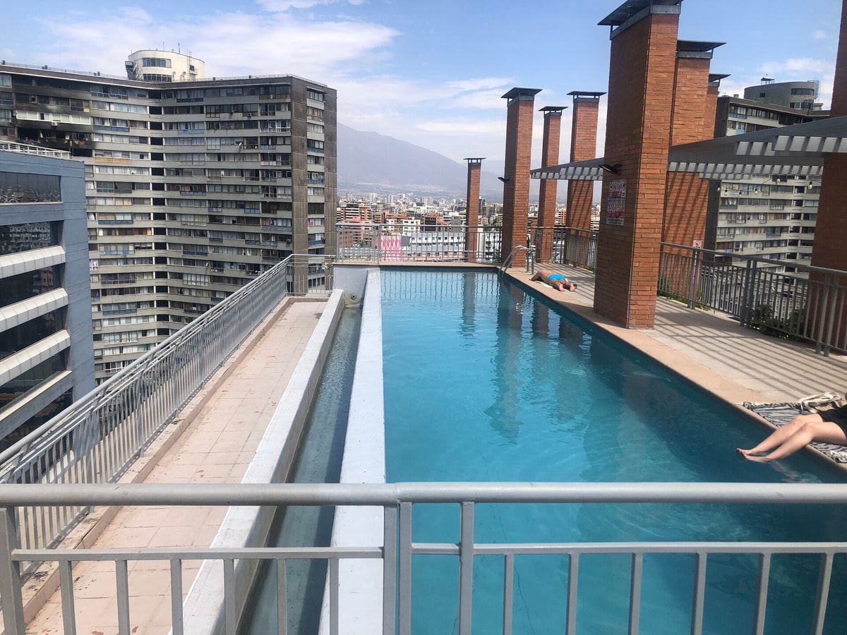Piscina del edificio en la azotea, larga y angosta, con solárium de baldosas y vista a los edificios del barrio y la cordillera