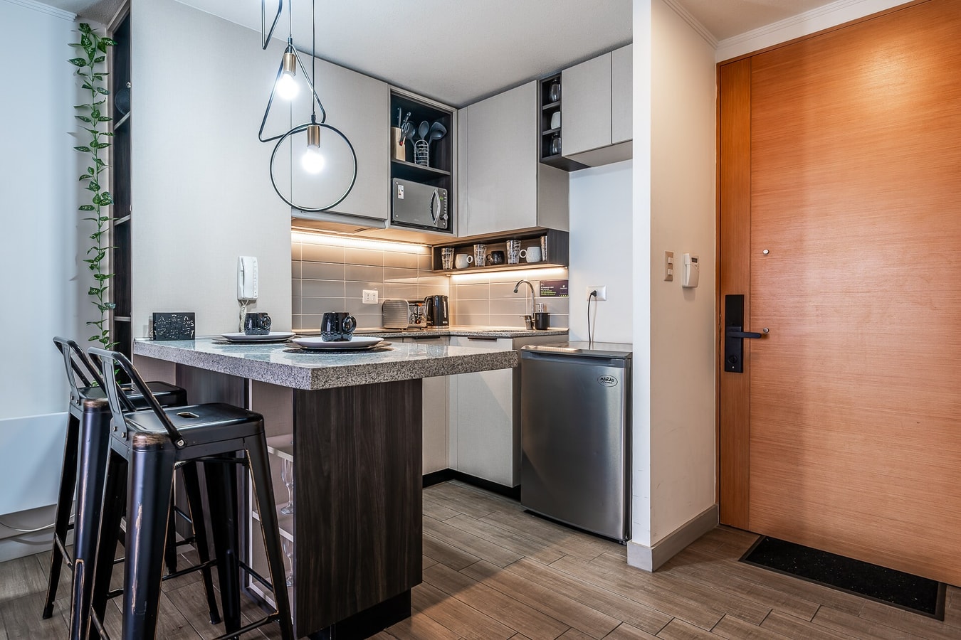 Cocina con barra y banquetas altas junto a la puerta de acceso de madera del departamento