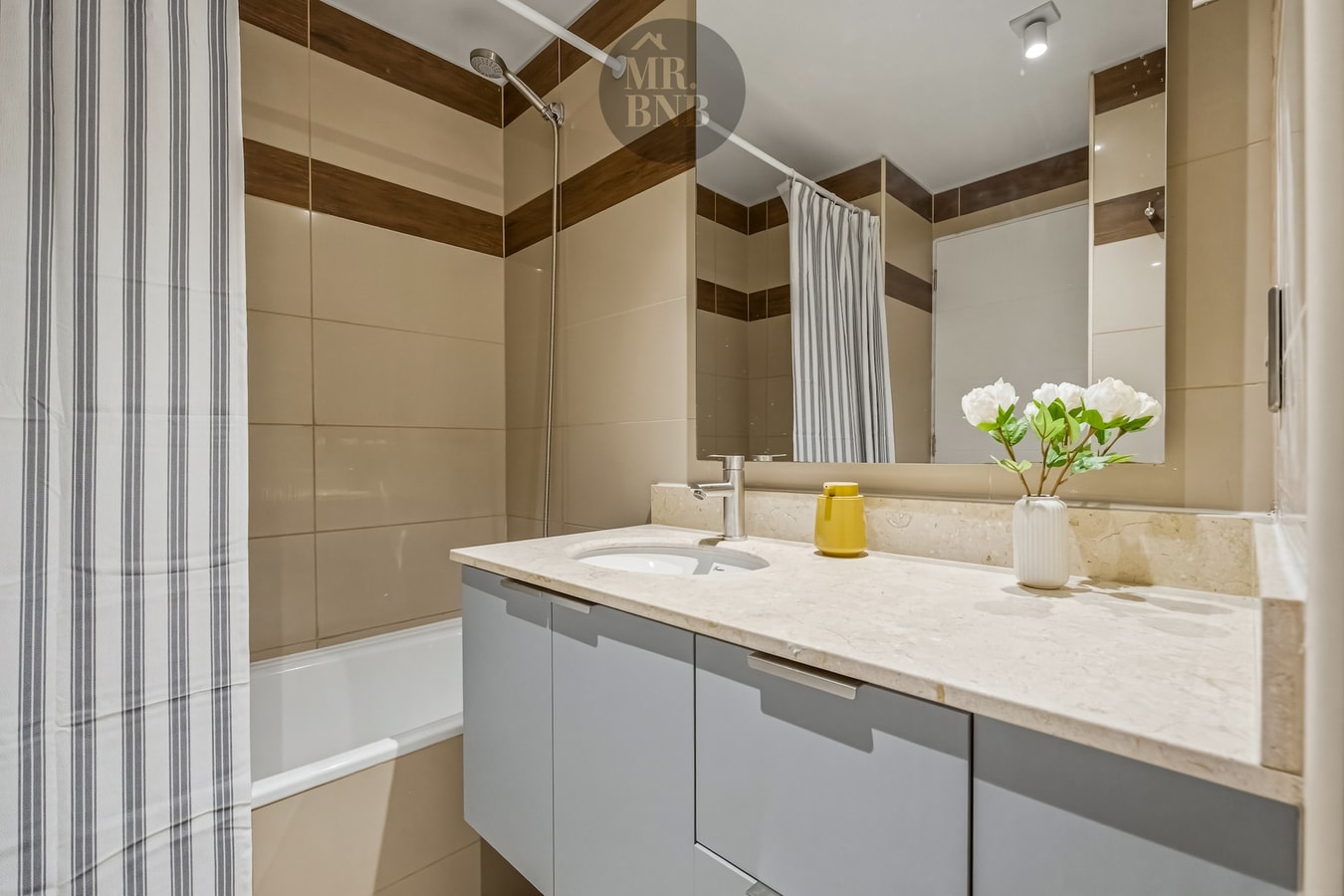 Baño con mueble vanitorio, espejo, flores sobre la encimera y tina con cortina a rayas