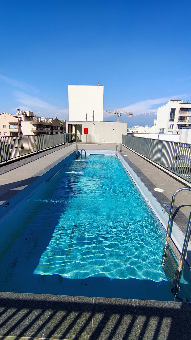 Piscina del edificio en la azotea, larga y angosta, con baranda perimetral y vista a los techos del barrio