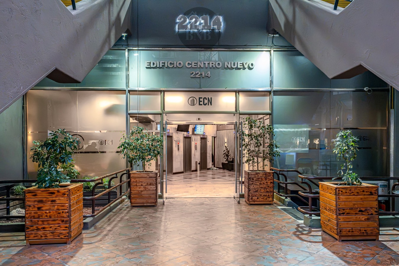 Acceso al edificio con hall iluminado, jardineras y letrero del edificio.