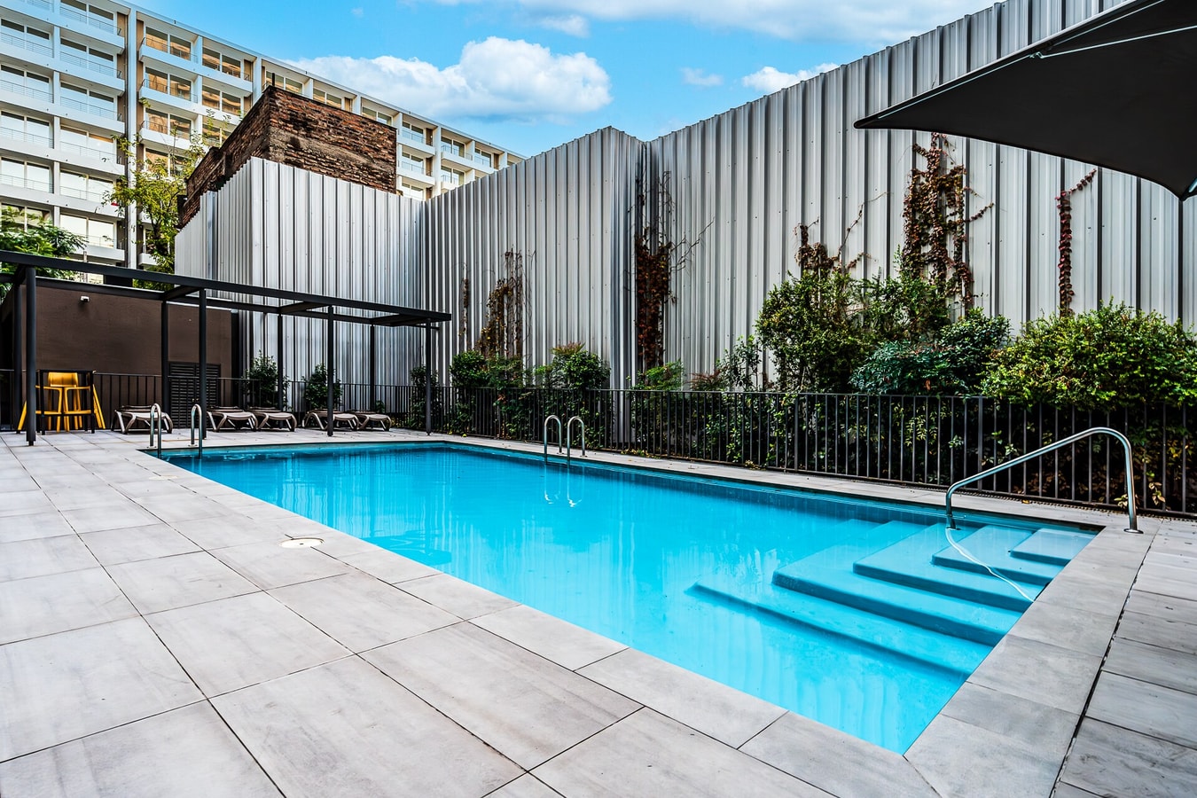 Piscina exterior del edificio, con solárium de baldosas, reposeras bajo quitasol y muro perimetral